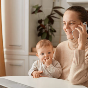 Indoor shot of happy smiling woman wearing beige casual style sweater with ponytail hairstyle sitting at table with her toddler daughter and talking phone with optimistic facial expression.
